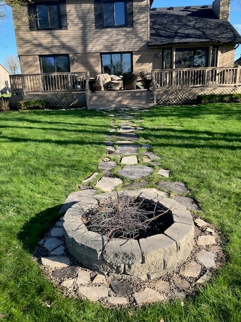 Stone fire pit with stepping stones leading through manicured lawn to brick house with wooden decks