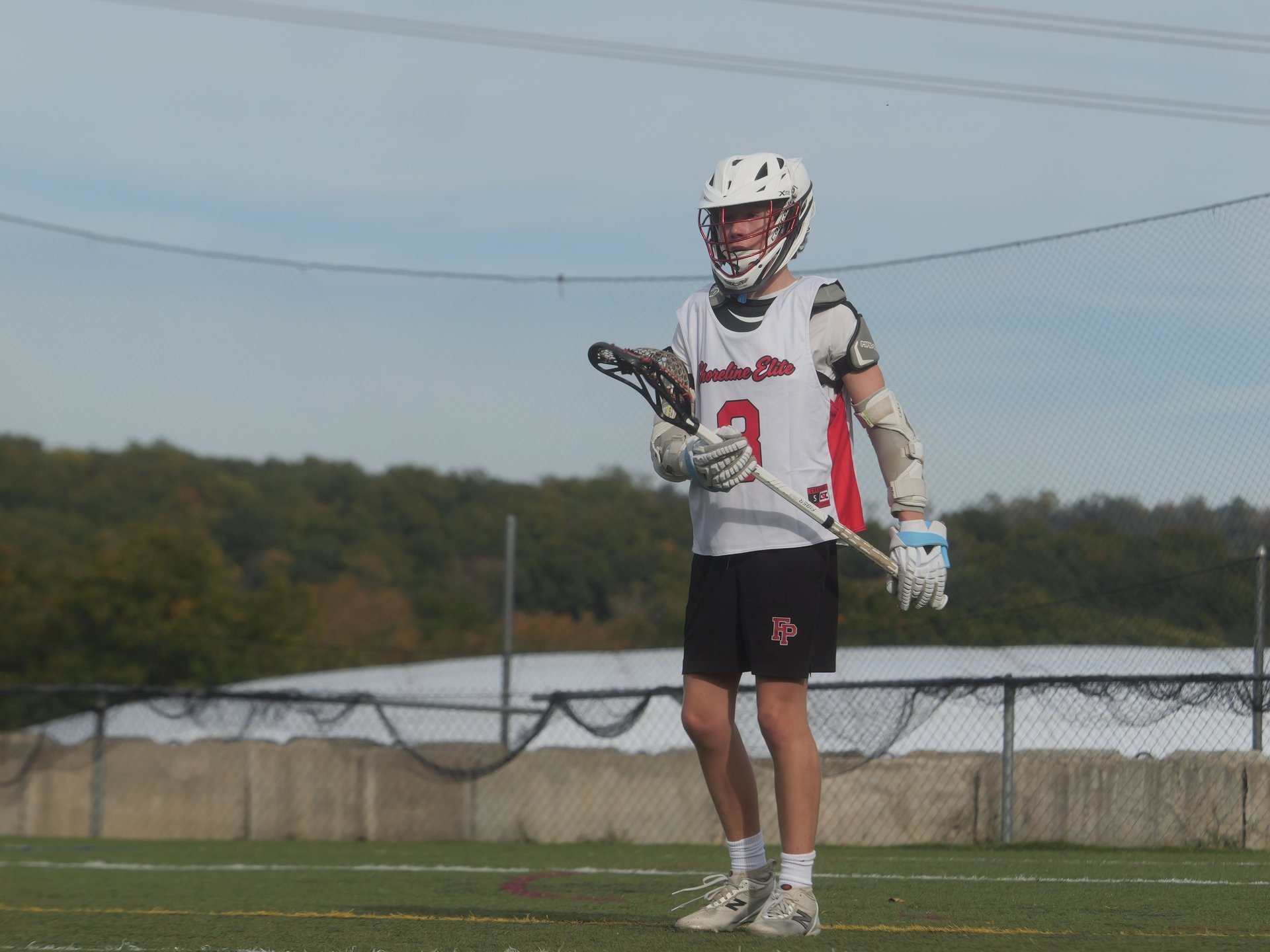 Lacrosse player in white and red uniform holding stick, standing on field with chain-link fence in background
