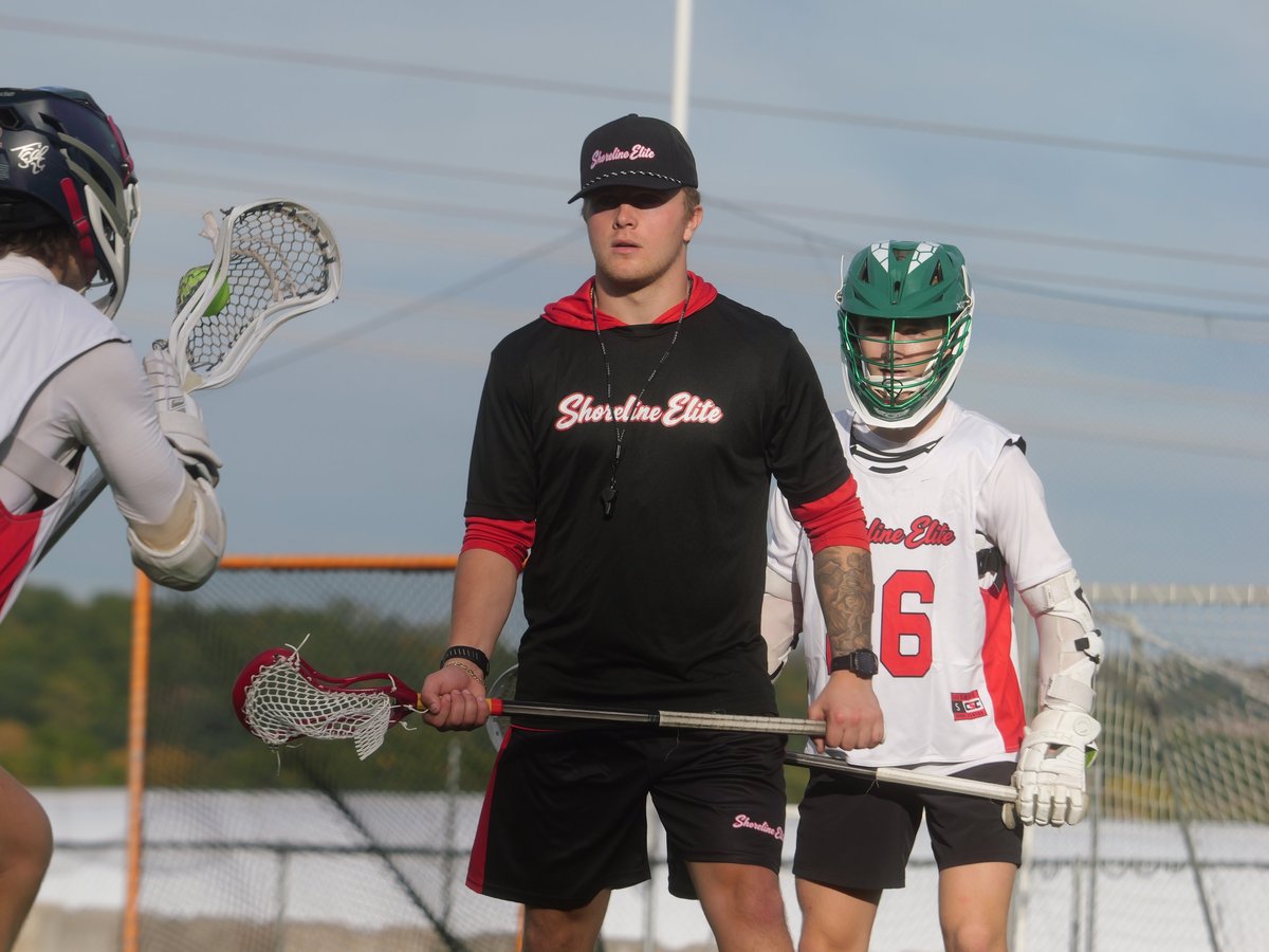 Shoreline Elite coach in black gear instructing two young players holding lacrosse sticks on an outdoor field