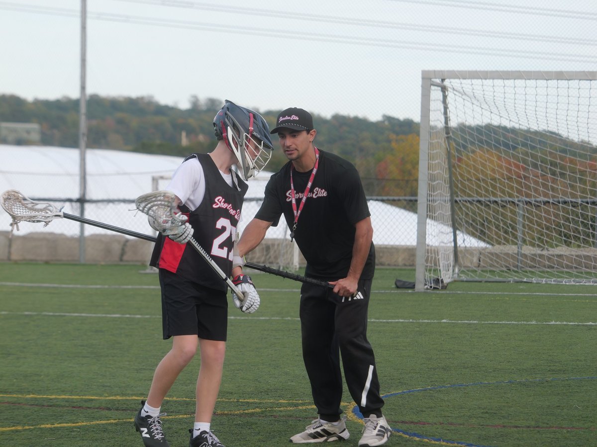Shoreline Elite coach instructing a young player holding a lacrosse stick on the field with the goal in the background
