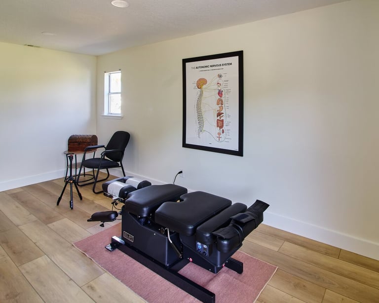 A chiropractic treatment room with a black massage table on a pink mat, seating area, and anatomical poster on the wall.