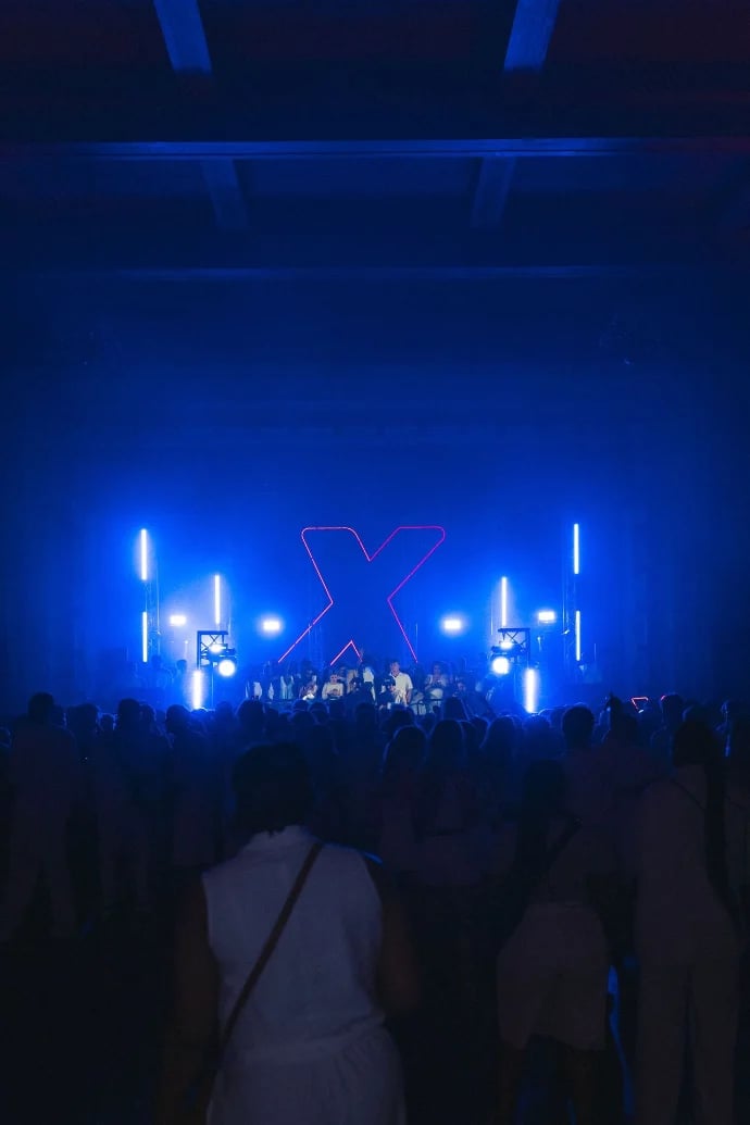 Concert stage with large illuminated X sign surrounded by blue neon lights and silhouetted crowd in foreground