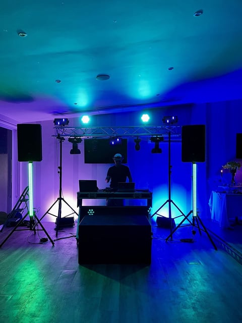 DJ setup with blue and purple stage lighting, speakers on tripods, and equipment on a table in a dark venue