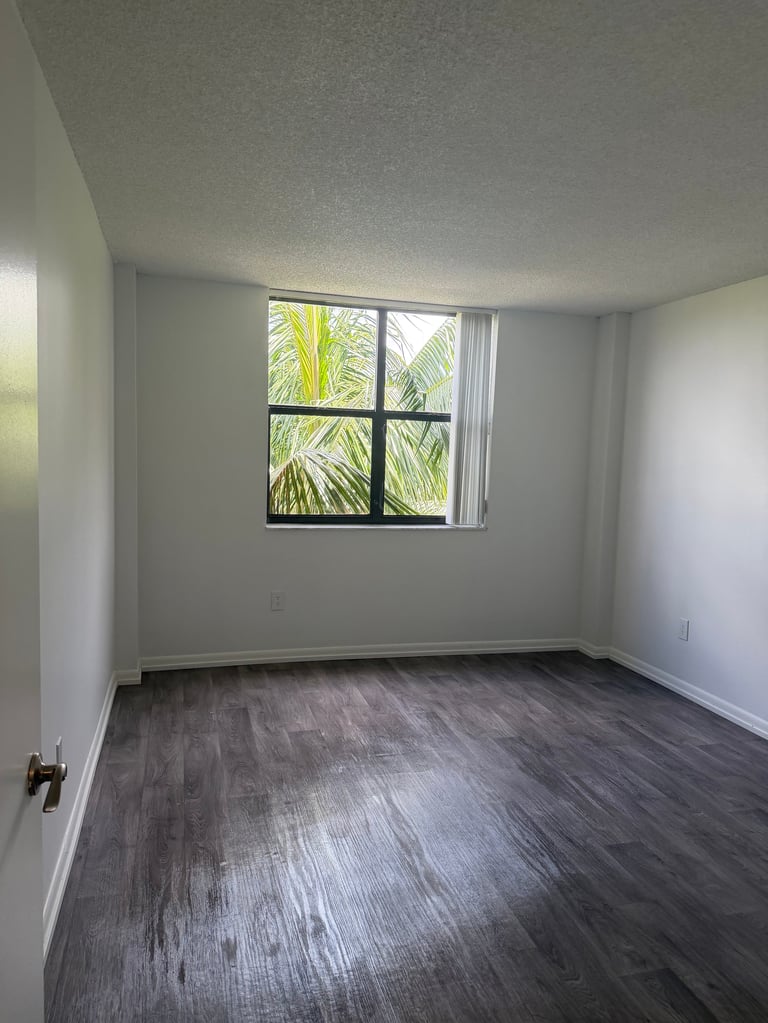 Empty room with gray walls, dark wood flooring, and a window showing palm trees outside