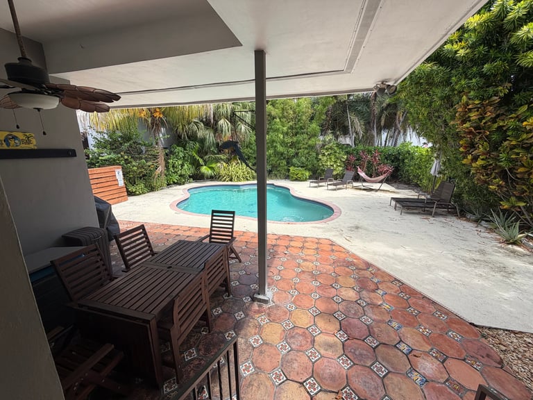 Covered patio with turquoise pool, wooden lounge chairs, and lush green foliage surrounding a brick terrace