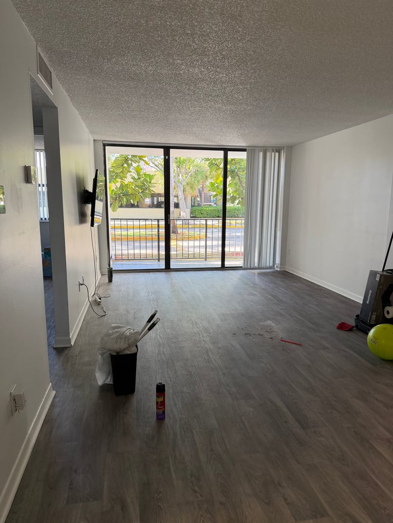 Empty apartment interior with dark wood flooring, white walls, and large sliding glass doors overlooking a green outdoor area