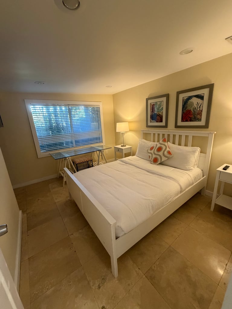 Bright bedroom with white bed, side table with lamp, large window with blinds, framed artwork on walls, and tiled floor