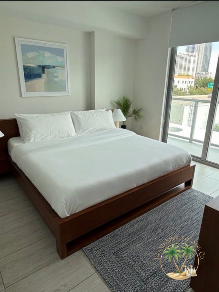 Modern bedroom with queen bed in dark wood frame, white bedding, ocean artwork, plant, blue area rug, and floor-to-ceiling window overlooking cityscape