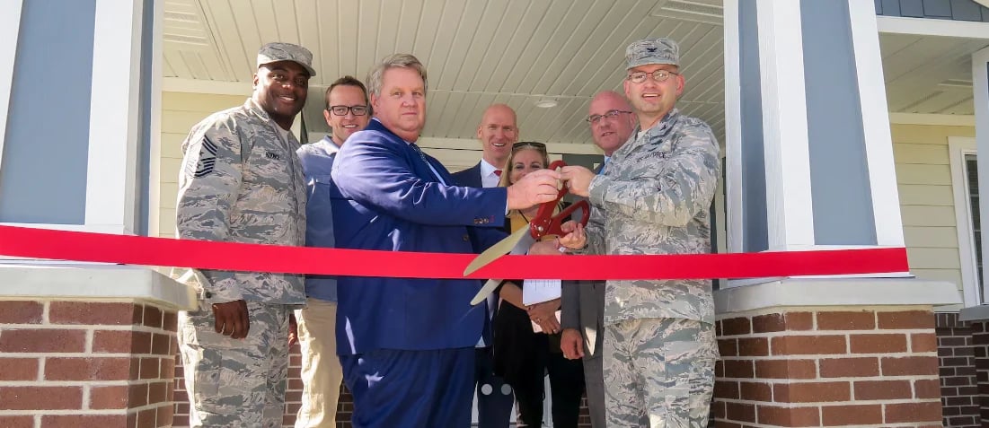 Group of people in military uniforms and civilian clothing cutting a red ribbon at a building dedication ceremony