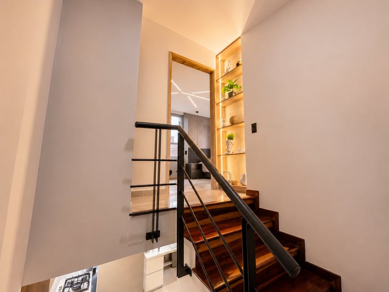 Modern staircase with wooden steps and black metal railing leading upward, with illuminated wall shelving and clean white walls