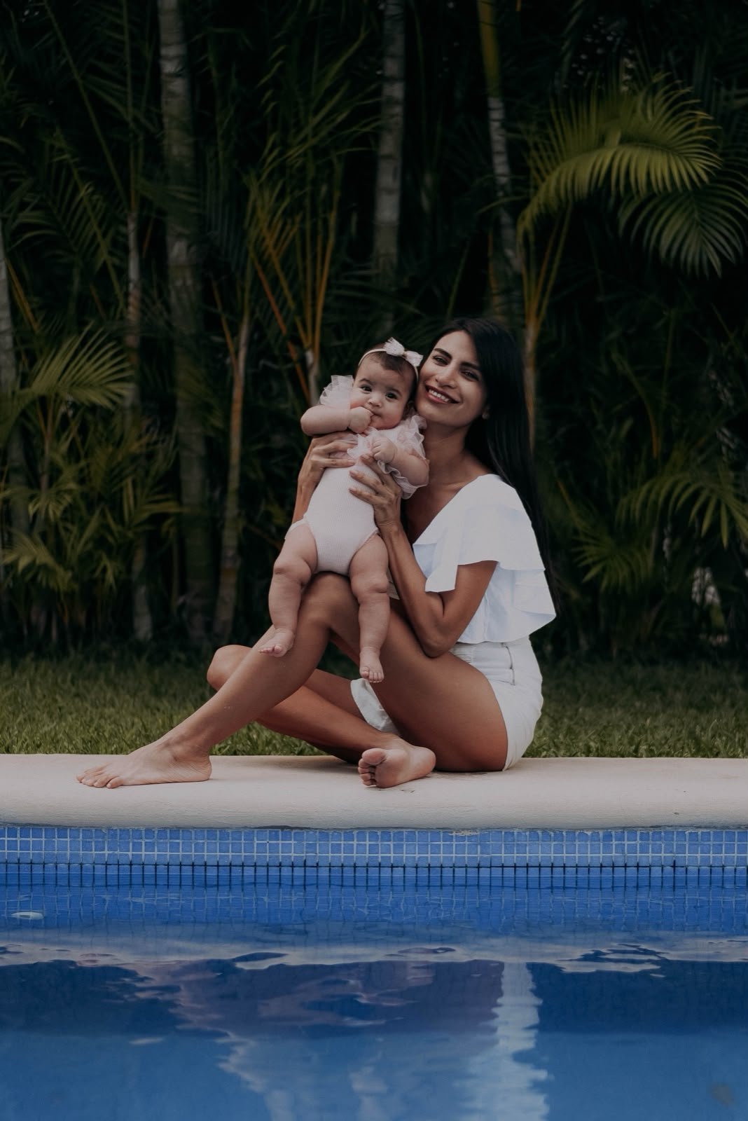 Mother and toddler in white clothing sitting by a swimming pool with palm trees in background