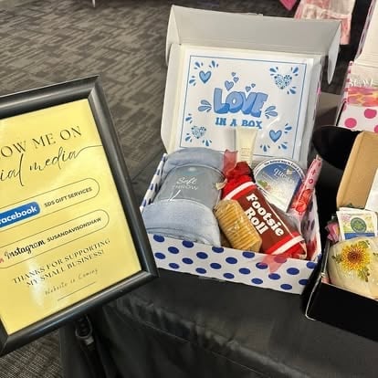 Display of gift boxes and items including a blue and white patterned box labeled Its a Boy, a yellow social media frame, and various small gift products on a table