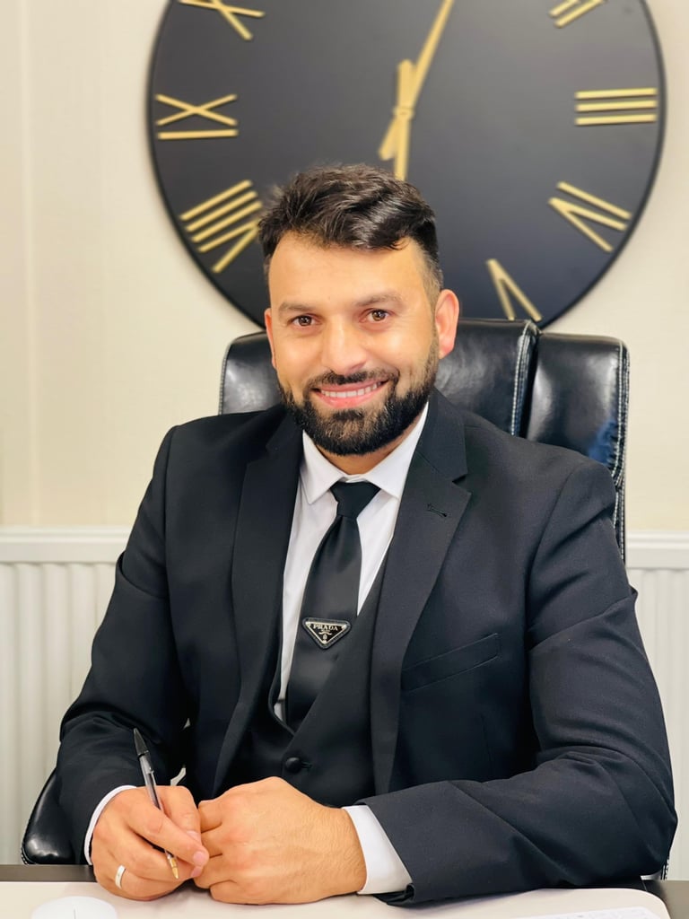 Syed Shakil Shah, solicitor at Legalise It Solicitors, in a black suit and tie sitting at his desk with a large wall clock in the background