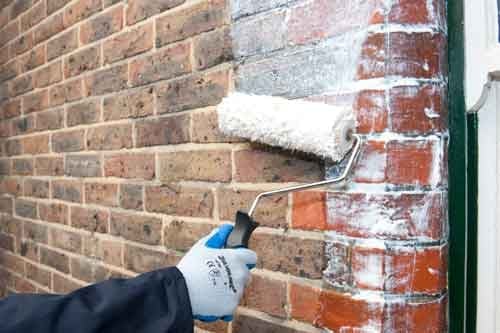 Worker in gloved hand painting weathered red brick wall with white paint using a brush roller