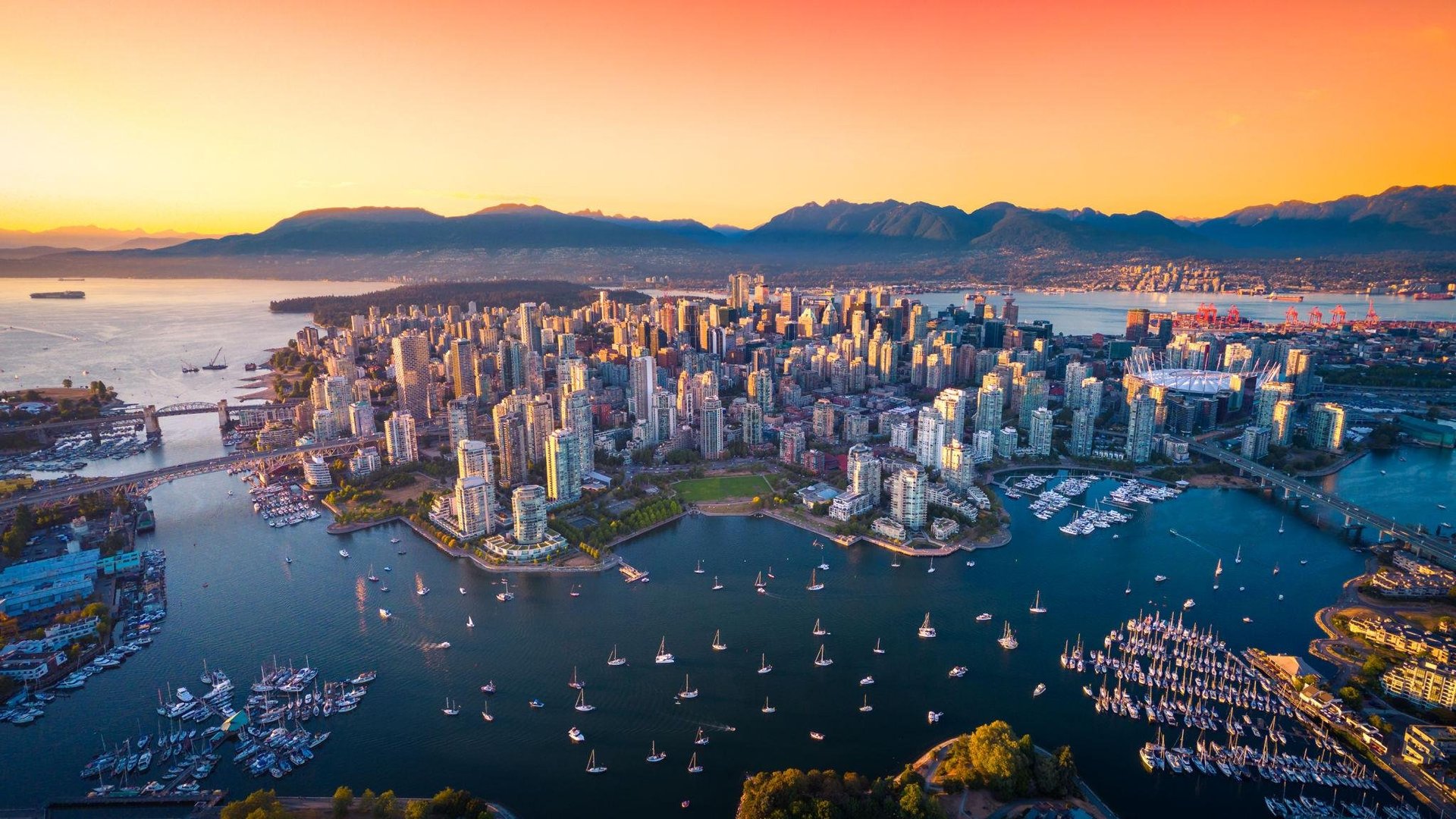 Aerial view of Vancouver at sunset with downtown skyline, harbor filled with sailboats, and mountains in the distance