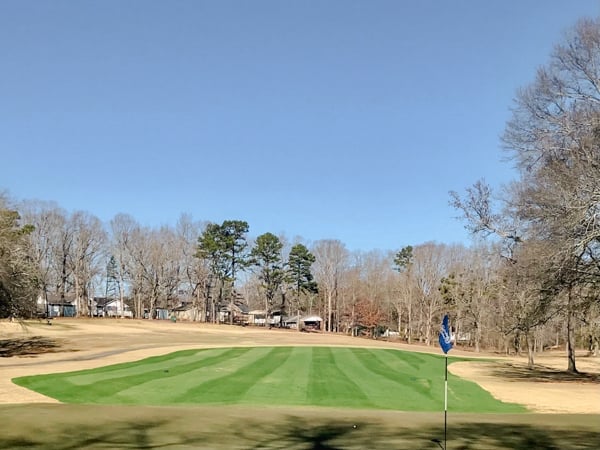 Well-maintained golf course with striped green fairway, flag pole, trees lining the course, and residential buildings in background on clear day