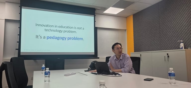 A man sitting at a conference table presenting, with a projector screen behind him displaying text about innovation in education.