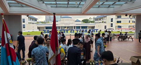 Students gathered in a school courtyard during an outdoor assembly or flag ceremony, with a large red flag visible in the foreground.