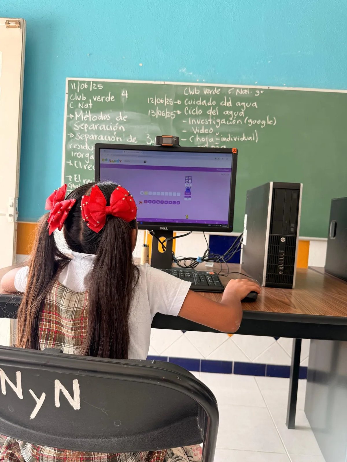 Student with long dark hair wearing a red bow sits at a desk in front of a monitor displaying a retro-style computer game in a classroom.