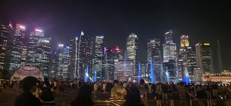 Crowded waterfront at night with illuminated skyscrapers and blue light installations reflecting on water