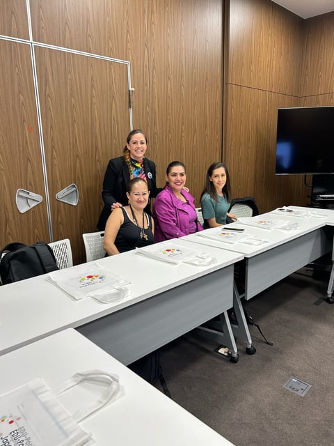 Four women posing together at a long white table in a wood-paneled conference room with papers and materials laid out