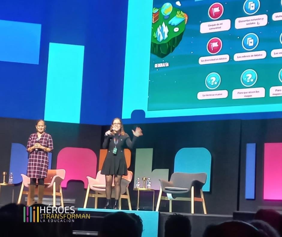 Two speakers presenting on stage with colorful chairs and a large screen displaying an educational game interface behind them.