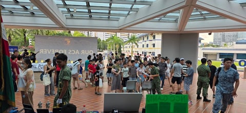 Large crowd of diverse people gathered in a modern indoor venue with wooden beams and skylights, appearing to be at a public event or expo
