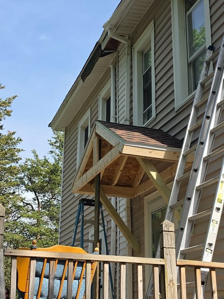 Wooden deck with railing attached to two-story house undergoing renovation with exposed roof framing and construction ladder
