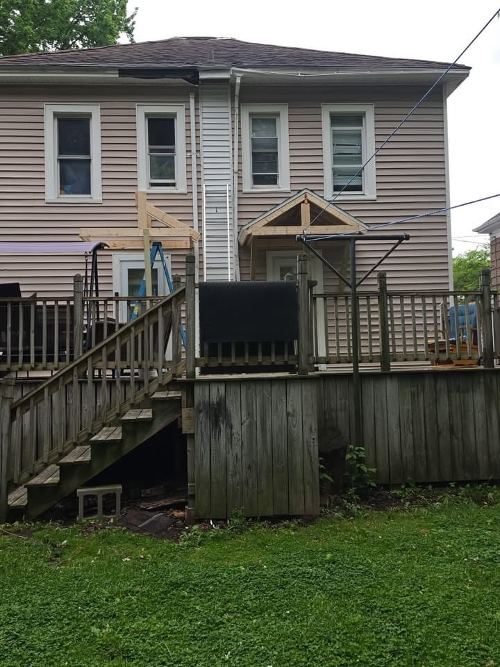 Two-story elevated house with gray siding, dark roof, wooden deck and stairs, green lawn, and surrounding trees