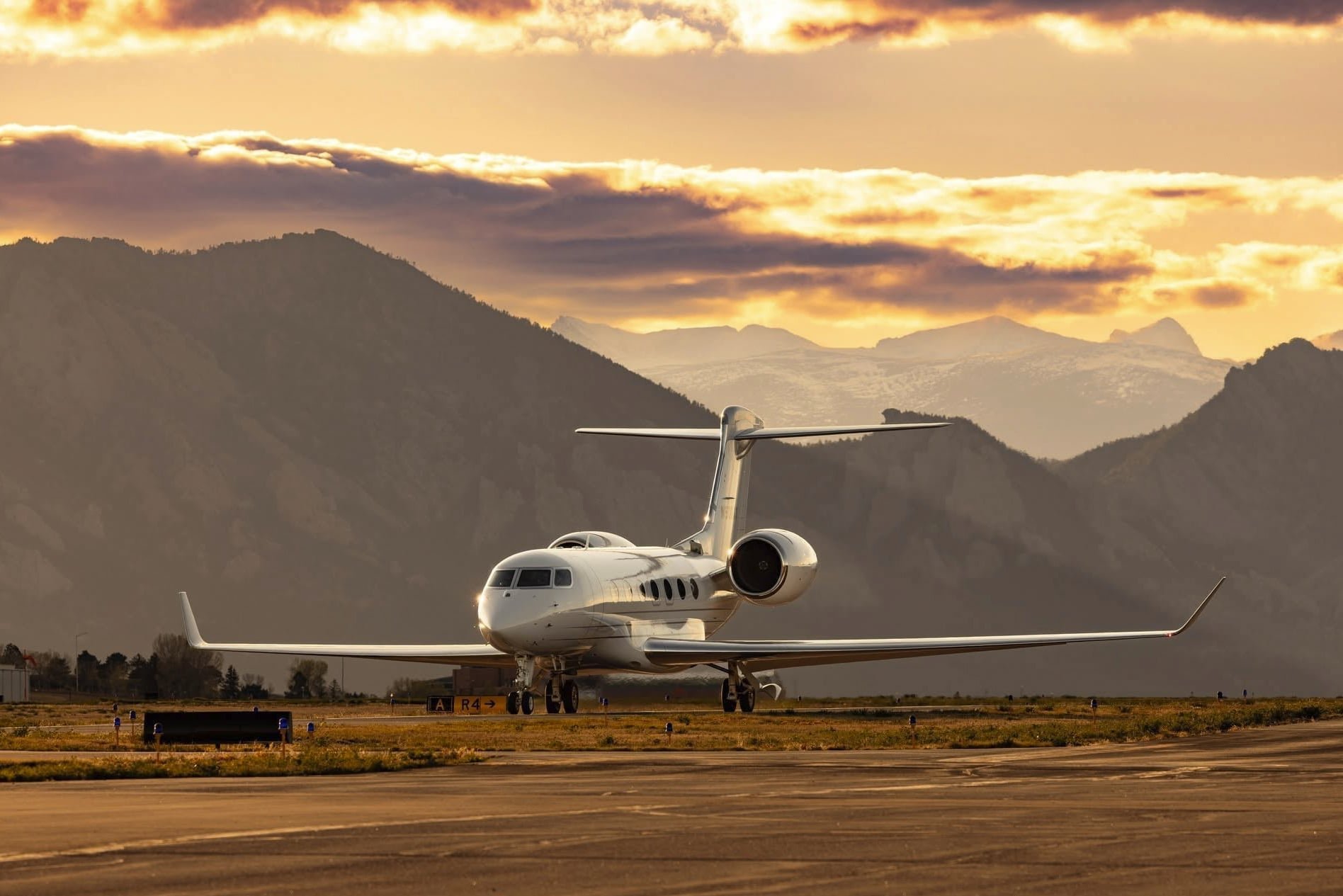 White private jet parked on runway with mountain range and dramatic sunset sky in background