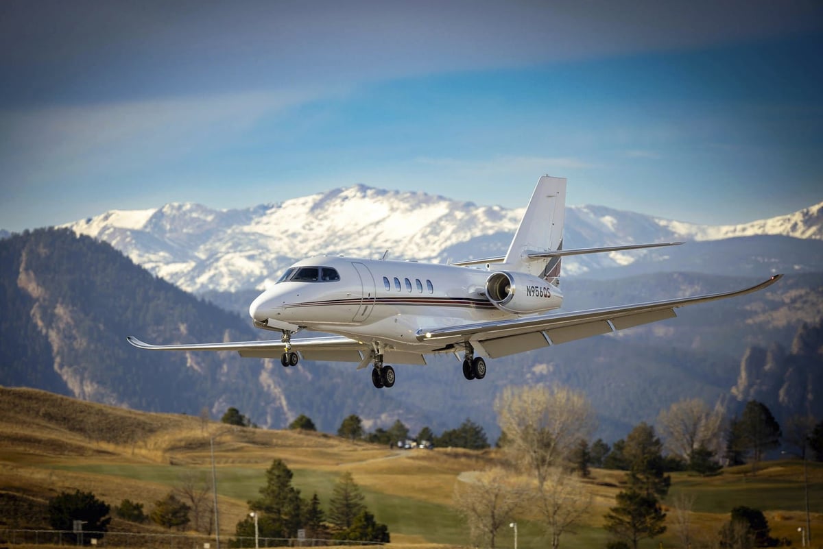 White luxury jet aircraft landing over mountain valley with snowy peaks and green meadows in background
