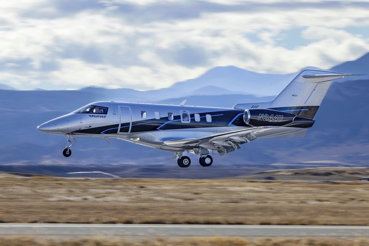 Blue and white business jet aircraft landing on an airfield with mountains visible in the background under cloudy skies