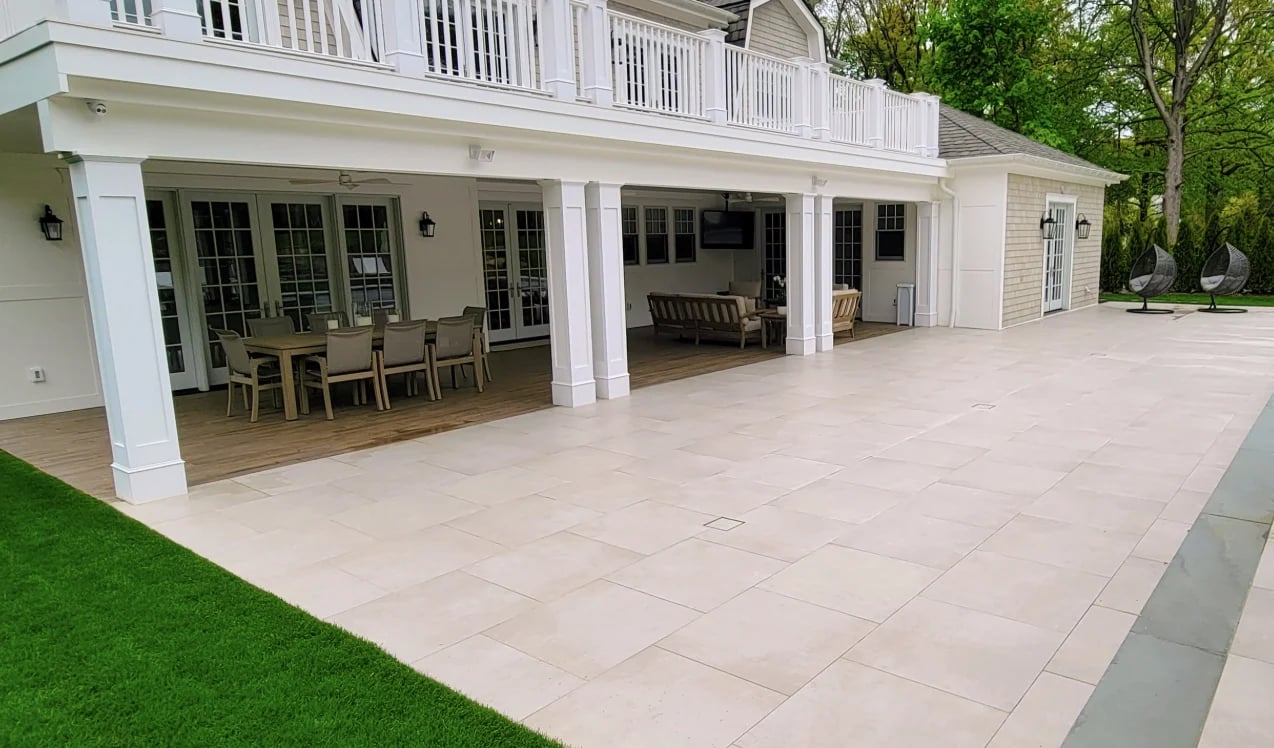 Modern white two-story house with open patio area, dining furniture visible through glass doors, surrounded by manicured lawn and trees.