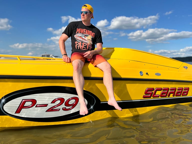 Man in yellow cap and black t-shirt sitting on yellow speedboat labeled P-29 in shallow water under blue sky
