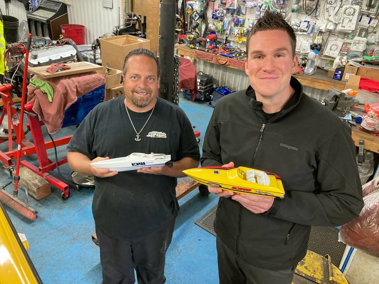 Two men in a workshop holding a yellow RC model airplane, surrounded by bikes and hobby equipment
