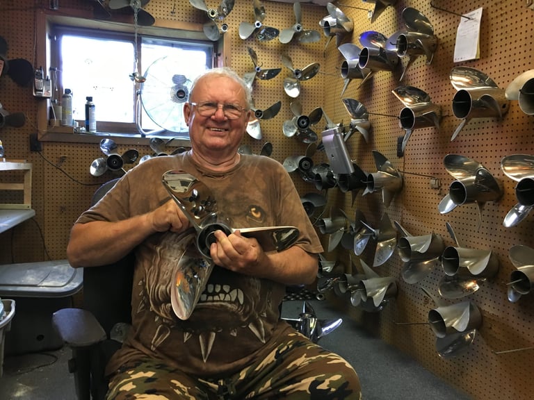 Man in a workshop holding a polished stainless steel propeller, surrounded by a pegboard wall lined with dozens of props and outdrives
