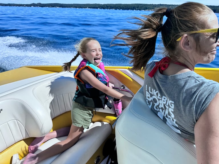 Two children laughing at the camera while riding on a yellow speedboat, both wearing life jackets and sunglasses with the lake wake behind them