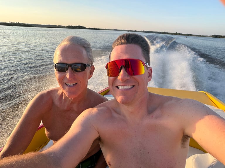 Two men on a speedboat wearing sunglasses, smiling at the camera with a wake visible behind them on the water