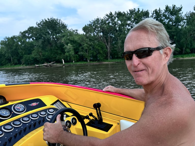Shirtless man wearing sunglasses operating a yellow and pink speedboat on a river with trees in the background