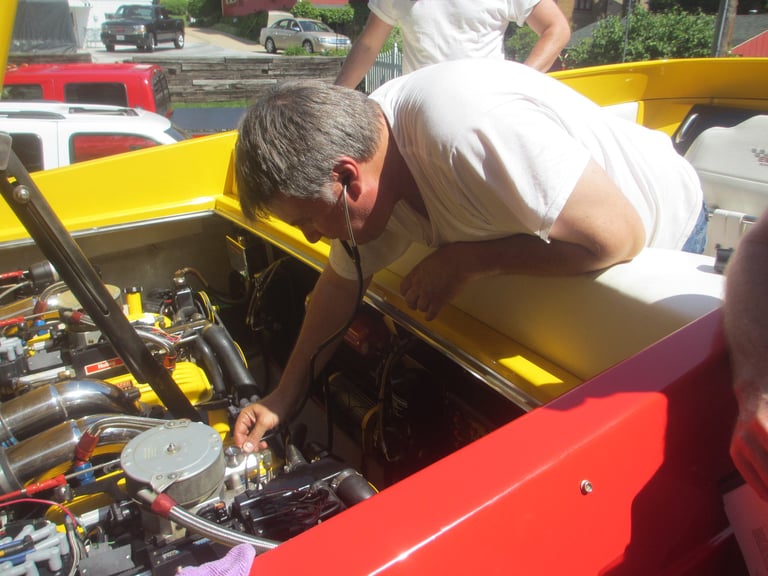 Man in white shirt leaning over yellow and red vintage car engine at outdoor car show