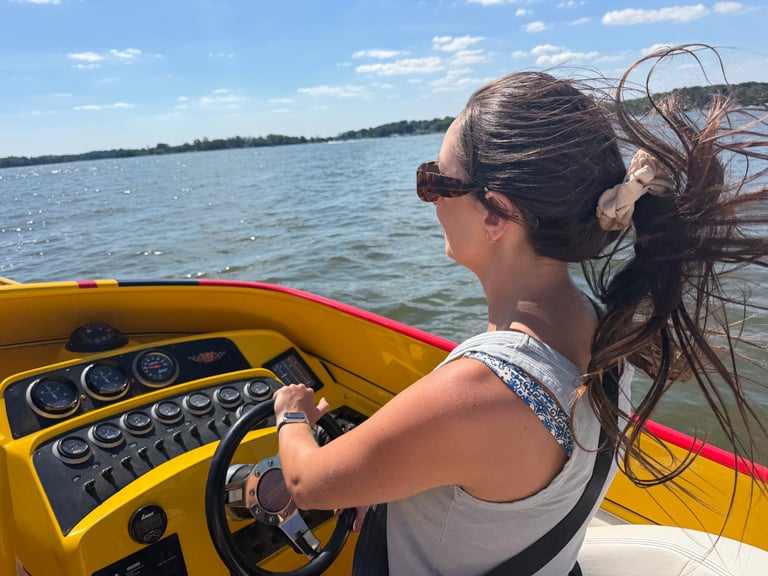Woman with brown hair in ponytail and sunglasses piloting the yellow and red P-29 across blue water, cockpit gauges and steering wheel in the foreground