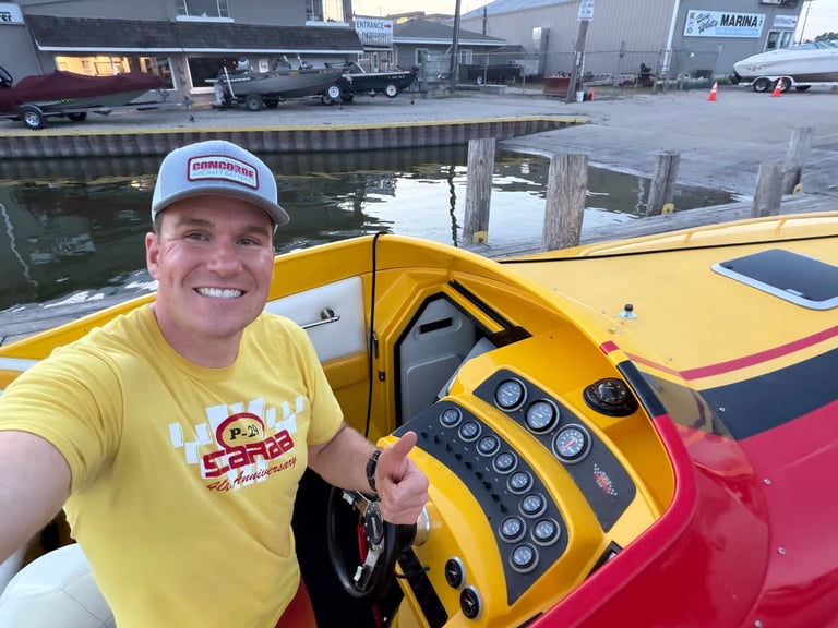 Man in a yellow Scarab 40th Anniversary t-shirt and Concorde cap smiling from the helm of the P-29 at a wooden dock with the rest of the dashboard visible beside him