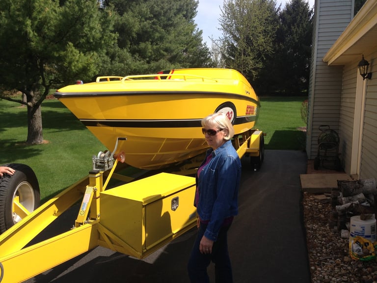 Woman in denim jacket standing beside bright yellow boat on trailer in residential driveway