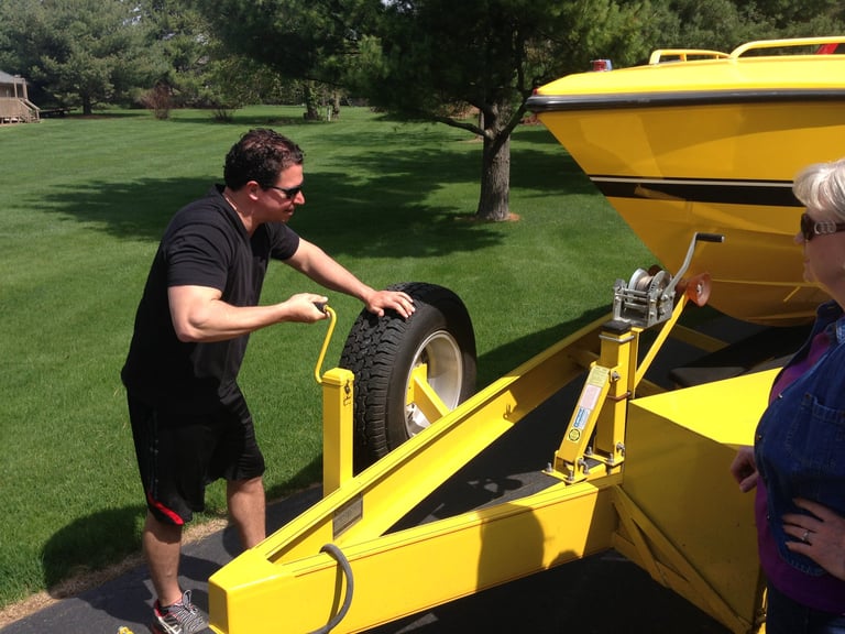 Man loading a spare tire onto the tongue of the yellow P-29 trailer in a front yard, with a woman watching