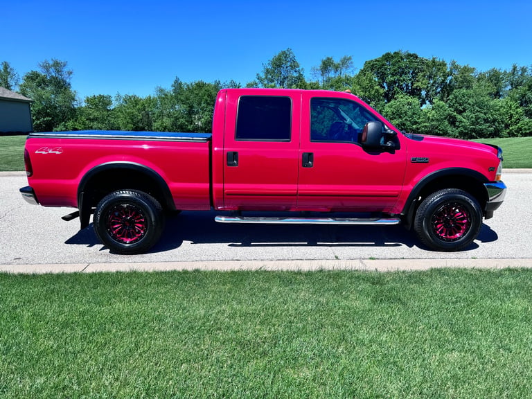 Bright red 2003 Ford F-250 extended cab pickup with black 4x4 off-road wheels and chrome side steps, parked on grass with blue sky and trees behind