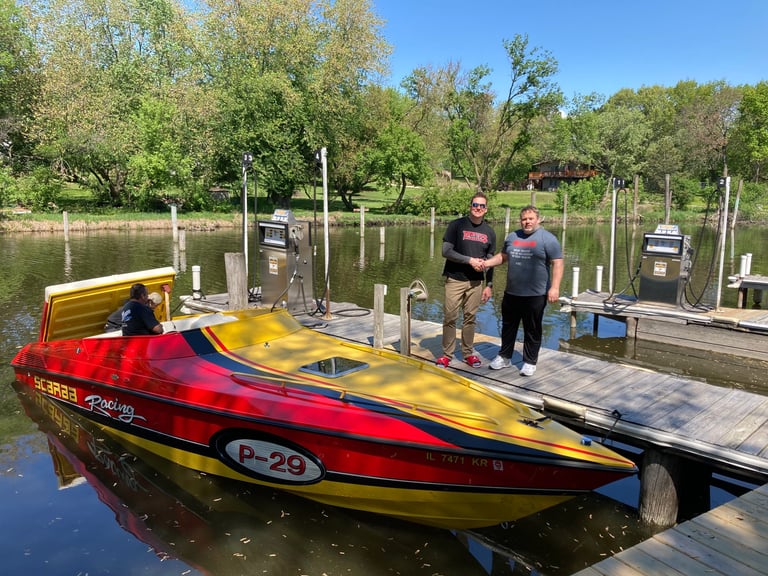 Red and yellow speedboat docked at a wooden pier with two men standing on the dock, trees and calm water in background