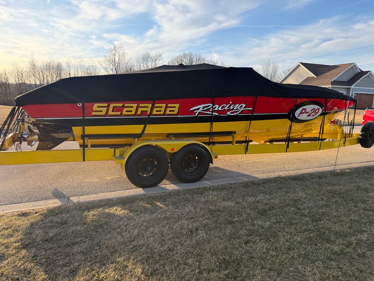 The P-29 under its black mooring cover on the yellow Scarab Racing trailer at dusk, parked in a driveway with the house and bare trees behind