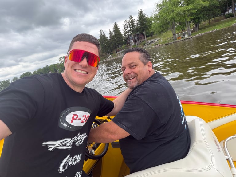 Two men smiling on a yellow and white speedboat on a lake with forested shoreline in background