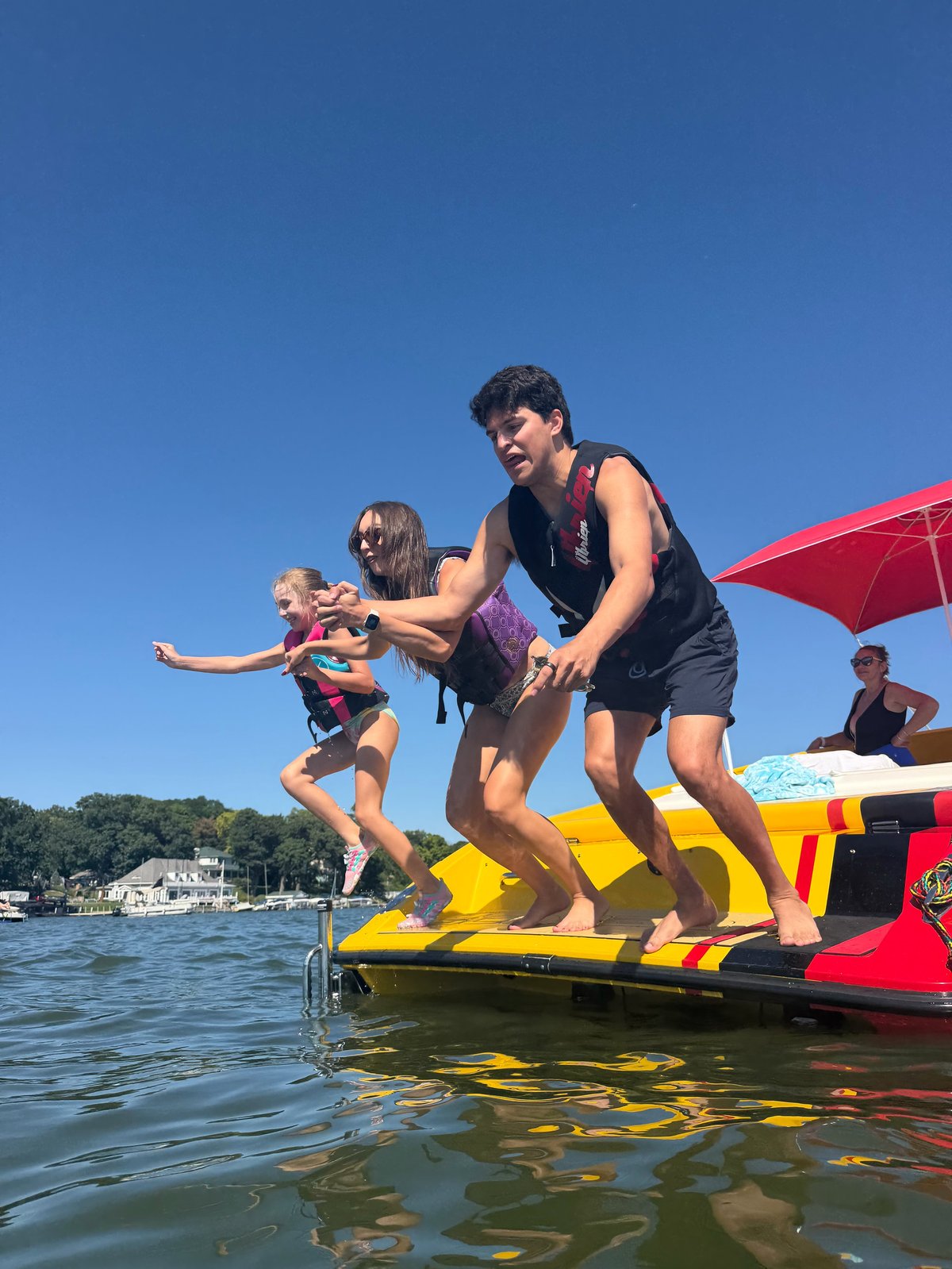 An adult helps children jump off a red and yellow boat into blue water on a sunny day