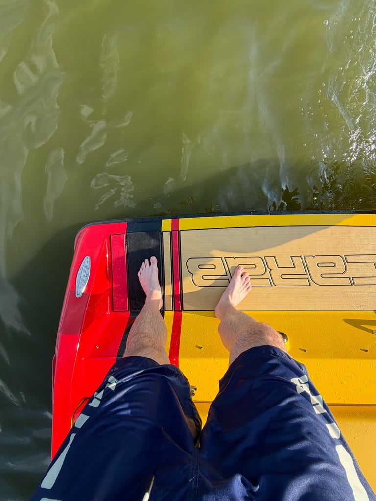Looking down over the transom from the swim platform, a barefoot owner standing on the SeaDek with the large SCARAB wordmark, red and yellow hull against green water